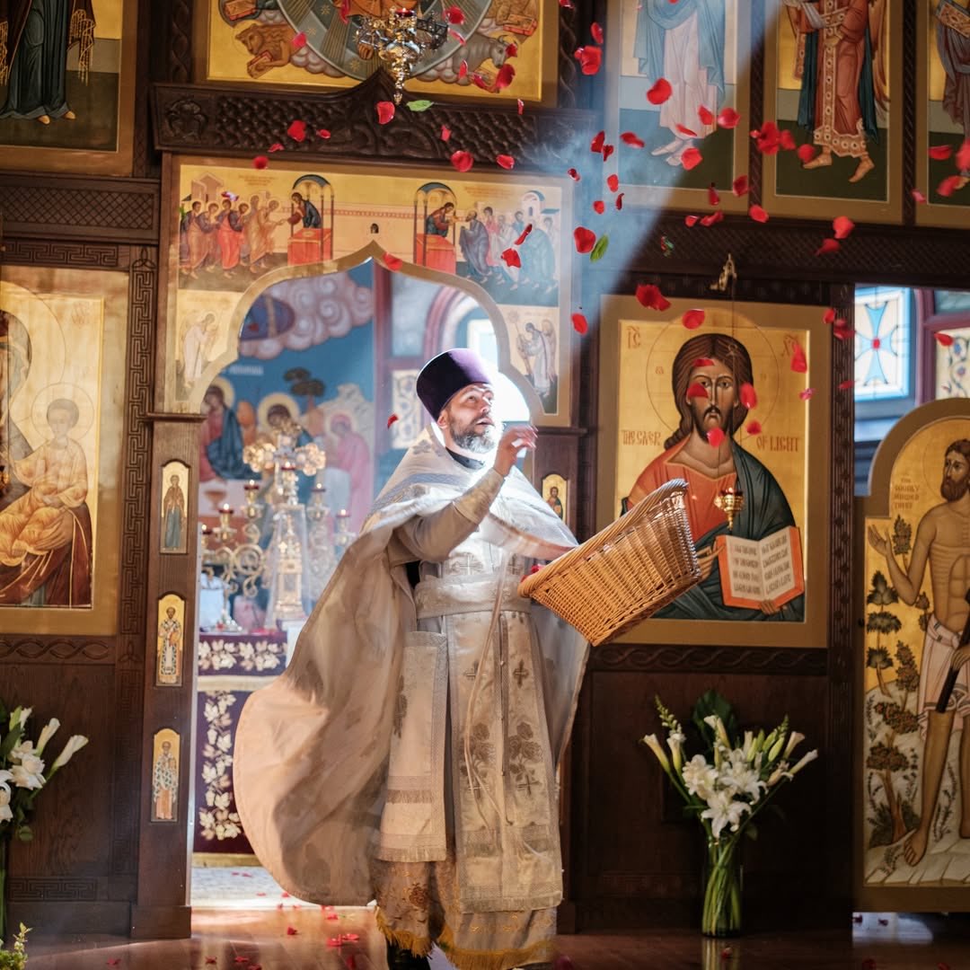 A priest in white and gold vestments scattering rose petals from a wicker basket before the iconostasis, petals falling through the air
